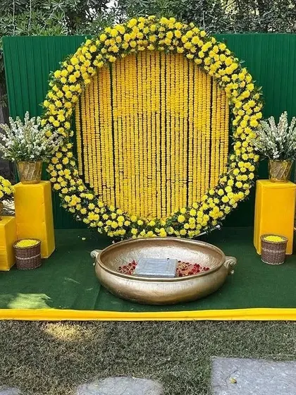 A wide view of an outdoor Haldi setup, showing the circular marigold backdrop, the 'urli' for the ceremony, and the surrounding lawn space.