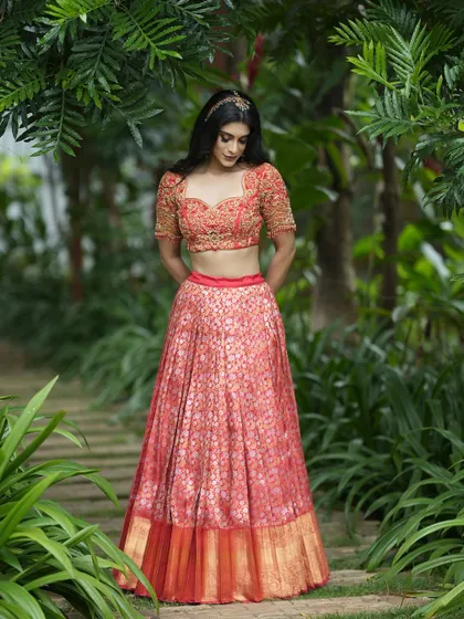 A signature style half saree featuring a coral-red brocade lehenga. The ensemble is paired with a heavily hand-embroidered blouse and a tissue-embroidered dupatta for added elegance.