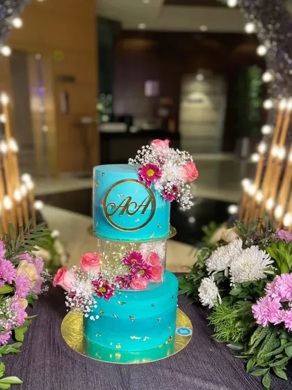 A beautiful two-tier engagement cake in a serene blue, with a floating middle tier. The cake is decorated with fresh flowers and a custom monogram, set against a backdrop of romantic lights.