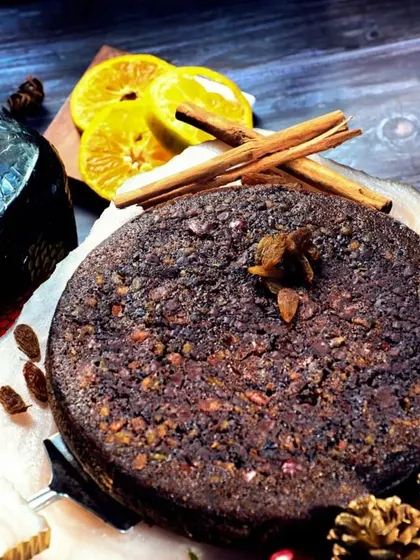 A dark and moody shot of a rich fruit cake, styled with cinnamon sticks and orange slices to evoke a warm, festive feeling.