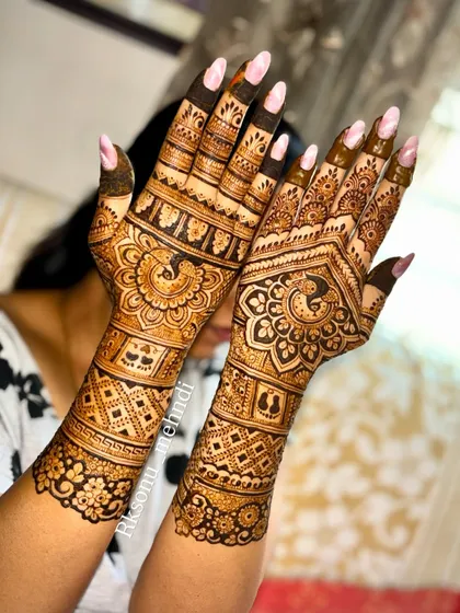 Full-arm bridal mehndi with a focus on peacock motifs. The design features bold, mirrored peacocks on each hand, surrounded by intricate check-work and floral bands that extend up the arms.