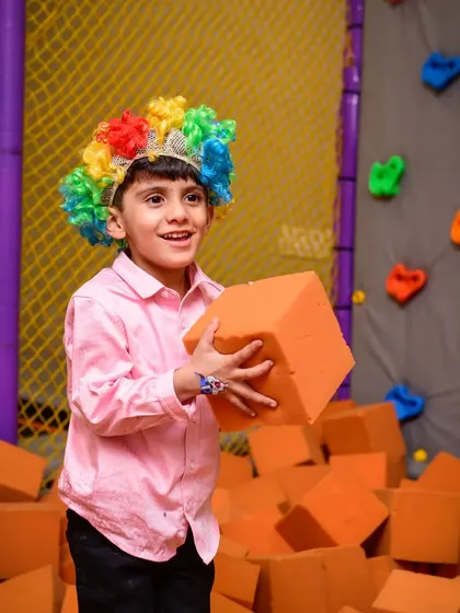 A boy in a colorful clown wig holds a foam block, ready to build something amazing in the foam pit.