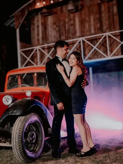 A stylish couple poses next to a vintage red car, with colored smoke adding a touch of modern drama to the rustic night setting. This showcases our creative use of props and effects for a unique pre-wedding photoshoot.