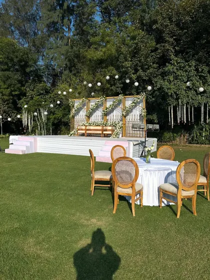 A wide view of the outdoor daytime stage. The white and gold structure, adorned with white roses, looks fresh and elegant on the green lawn.