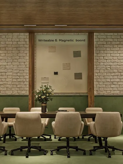 This meeting room combines formal structure with natural textures. A large wooden table is surrounded by comfortable swivel chairs, set against an exposed brick wall and a large, writable magnetic board for brainstorming sessions.