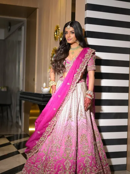 A full-length portrait against a modern backdrop. The bride looks like a contemporary princess in her pink lehenga, with makeup that enhances her natural beauty.