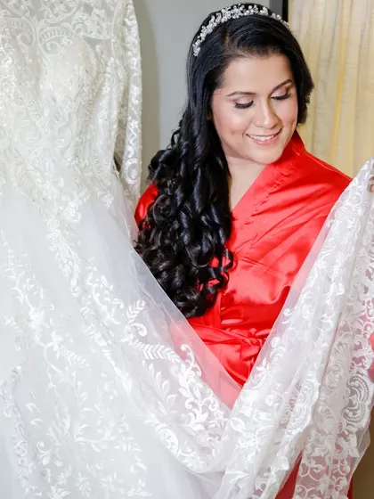 The bride-to-be in her robe, admiring the intricate lace of her wedding gown. Her hair and makeup are already done, showing the complete look before the final step.