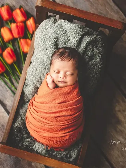 Blooming with love. This newborn is nestled in a tiny wooden bed, swaddled in a warm orange wrap next to fresh tulips, creating a beautiful, artistic portrait that feels like a dream.