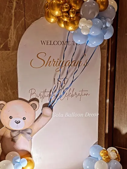 A personalized welcome sign for "Shriyan's 1st Birthday Celebration," featuring a cute teddy bear cutout holding a string of balloons.