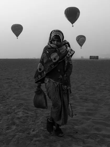 A woman walks across the sand as hot air balloons float in the sky during the Varanasi balloon festival. This black and white image creates a surreal, dreamlike scene.