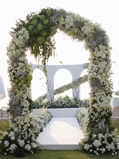 The full view of the floral archway leading to the ceremony space. The arch is dense with white and green florals, creating a grand and welcoming entrance for the bride and guests.