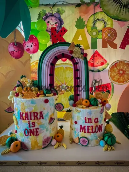 The cake is the centerpiece of the celebration. Here, two custom cakes reading "Kaira is ONE in a MELON" are perfectly integrated with the fruity backdrop and miniature fruit decorations.