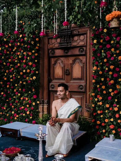 The groom awaits his bride, seated before a magnificent wall of vibrant flowers and a traditional carved door, a setting we designed to be both grand and personal.