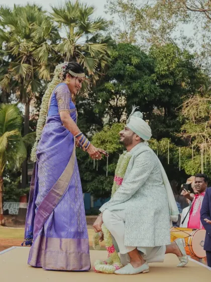 The groom kneels during the Varmala ceremony, a playful and romantic moment set against the backdrop of an outdoor celebration.