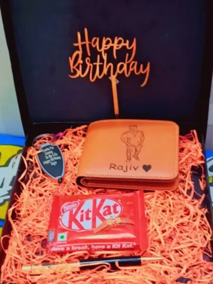 A simple and elegant birthday gift box containing a photo-engraved wallet, a personalized pen, and a keychain. The box is filled with festive orange shredding paper.