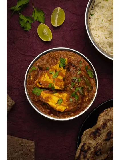 A rich bowl of paneer curry, styled with fresh roti and lemon wedges on a deep purple surface. The overhead lighting and vibrant colors make this dish look incredibly appetizing for a menu or food blog.
