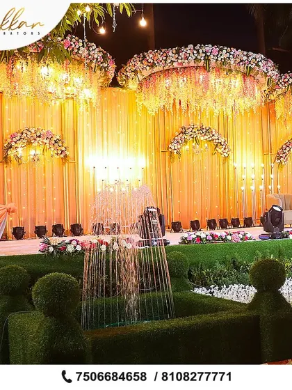 A grand reception stage with a unique fountain installation made of greenery. The backdrop of hanging flowers and warm lighting adds to the magical, garden-like feel.