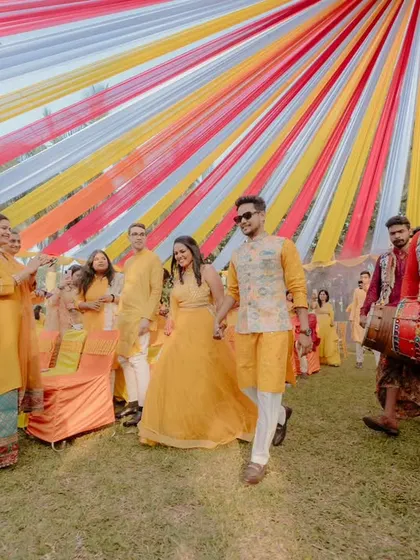 The groom's energetic entry with a dhol player, surrounded by family and friends dressed in yellow. The multi-coloured fabric canopy we installed provides a festive backdrop for this joyous procession.