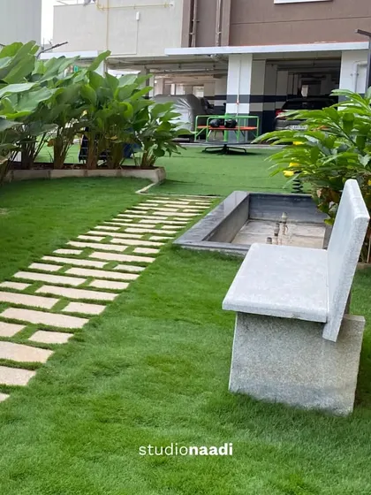 A simple granite bench provides a pause point along a garden path at Saritha Splendor. This functional element is integrated into the landscape, offering a place for quiet reflection amidst the greenery.