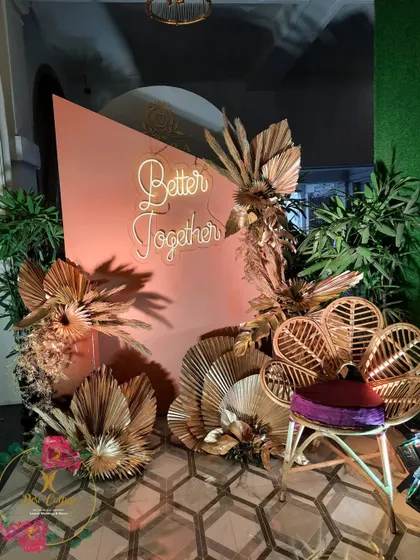 A stylish photo booth with a "Better Together" neon sign, a peacock chair, and arrangements of dried, gold-painted palm leaves.
