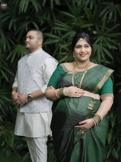 A lovely portrait focusing on the mother-to-be, with her partner in the background. The vibrant green saree stands out beautifully against the leafy backdrop.