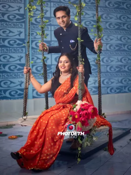 A sweet and traditional indoor pre-wedding photo on a decorated swing. The bride's orange saree adds a beautiful pop of color to the scene.