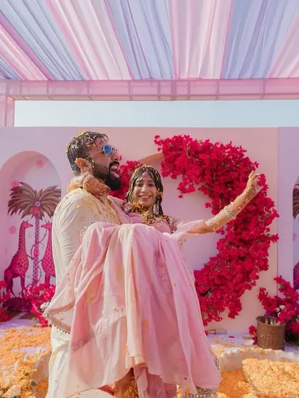 A playful moment at the end of a Haldi ceremony, with the groom carrying the bride against a custom-designed backdrop featuring pink giraffes and bougainvillea.