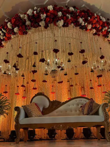 A beautiful reception seating area with a royal diwan set against a backdrop of cascading white and red florals and a crystal chandelier.
