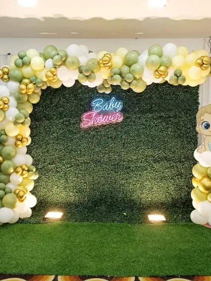 A simple and elegant baby shower photo booth. This design uses a green hedge wall as the backdrop, a 'Baby Shower' neon sign, and a balloon arch in muted green, white, and gold tones.