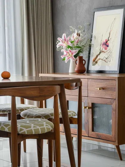 In our 'Poorna' project, the dining space exudes a timeless charm with its dark wood furniture set against a textured grey wall. The custom crockery unit with fluted glass doors adds a touch of vintage elegance.