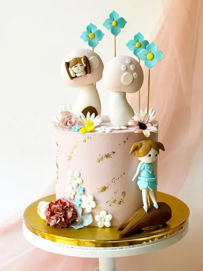 An enchanting woodland-themed cake. This pink cake is decorated with gold leaf, flowers, and adorable toppers including a girl figurine and whimsical mushrooms.