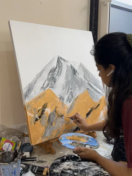 In my studio, working on a textured mountain painting. You can see my paint palette and the tools I use to create different effects on the canvas.