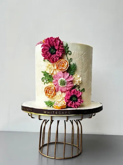 This tall celebration cake features intricate, hand-piped buttercream flowers in vibrant pinks and oranges, set against a subtly textured white frosting. It's a perfect example of the elegant, handcrafted wedding and anniversary centerpieces I love to create.