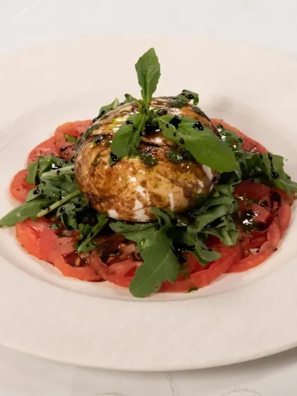 A fresh Burrata salad, served on a bed of sliced tomatoes with rocket leaves and a balsamic glaze. It is a creamy, fresh, and delicious starter.