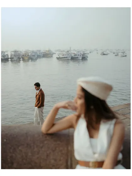 A creative shot using depth of field to tell a story. She is in soft focus in the foreground, looking thoughtfully away, while he stands by the water in the background, with the boats of Mumbai harbor behind him.