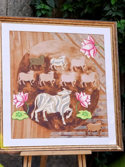 This framed artwork from the Nandi Collection is set against a natural backdrop, highlighting the earthy tones of the painting and the intricate embroidery of the sacred cows and lotuses.