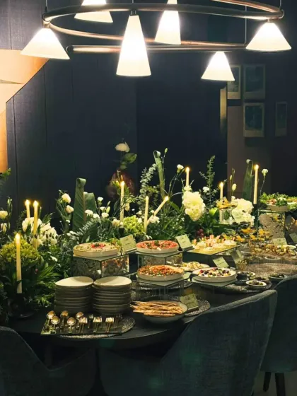 A long shot of a beautifully lit dining table transformed into a grazing station for an evening party. The setup includes multiple platters, pizzas, and salads, illuminated by candles and modern lighting for a chic, sophisticated ambiance.