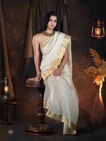 A full-length portrait showcasing a Kerala kasavu saree. The model is posed on a vintage stool amidst an earthy, rustic set, creating a timeless and artistic representation of traditional fashion.