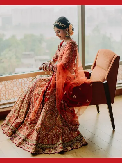 An elegant, full-length portrait of the bride, Maheswari, seated by a window, showcasing her regal poise and beautiful lehenga.