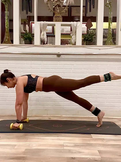 Pilates is about precision and control. Here, I am holding a plank with a leg lift, using ankle weights for added resistance and light dumbbells to engage my arms and shoulders. This single pose is a full-body exercise that strengthens the core, glutes, and upper body simultaneously.