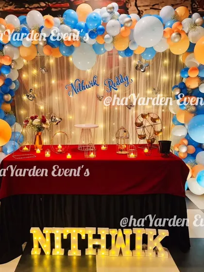 A full view of a table setup in front of a fairy light curtain, decorated with an orange and blue balloon arch.