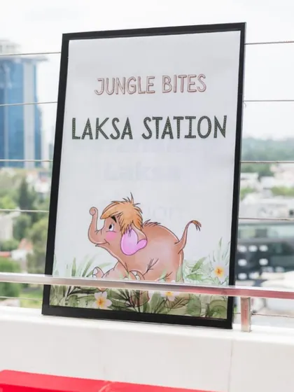 When rain changed our plans, we brought the jungle indoors. This sign for the "Jungle Bites Laksa Station" shows how we carry the theme through every detail, even the food service.