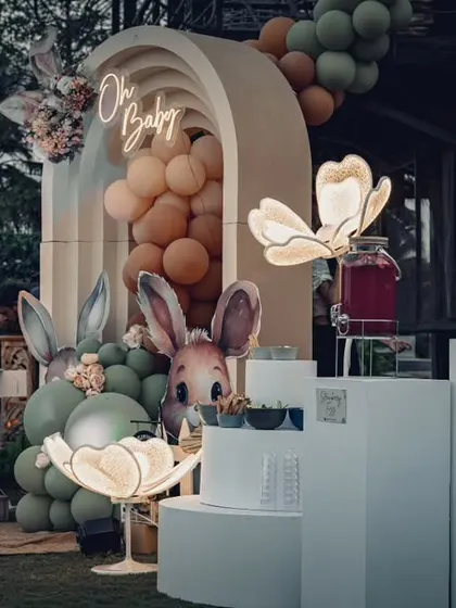 An outdoor "Bunny & Berries" themed baby shower at dusk. The glowing floral lamps and neon "Oh Baby" sign add a magical touch to the whimsical setup.