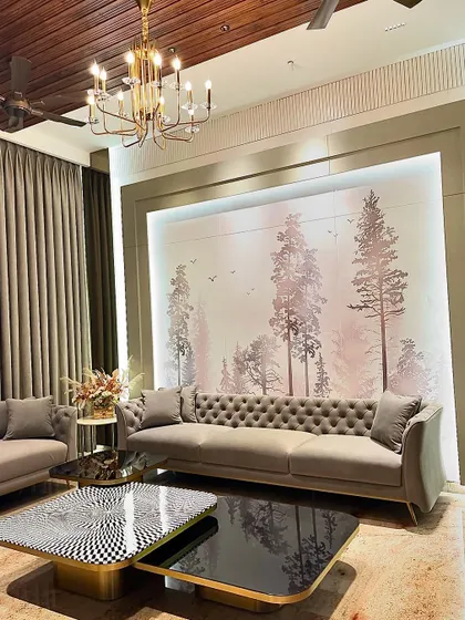 In this Ambala living room, a classic tufted sofa is set against a custom-designed wallpaper depicting a serene forest scene. The backlit panel creates a tranquil atmosphere, enhanced by the grand chandelier above.