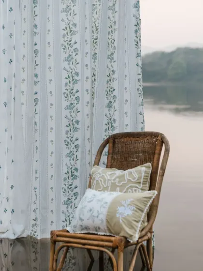 Our sheer curtains and embroidered cushions from the 'Pause' collection, set against a serene lakeside. The herb-green embroidery on the cushions complements the delicate prints on the drapes.