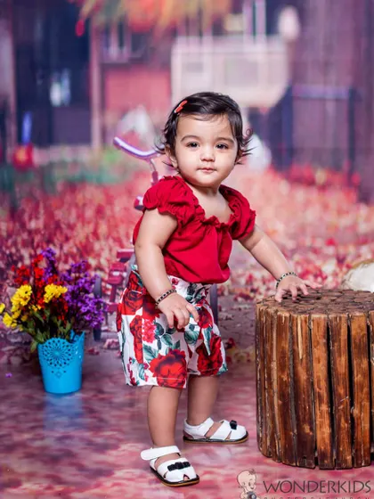 A toddler in a stylish red and white floral outfit, posing against a beautiful autumn-themed backdrop with red leaves. Her confident look and chic clothes make for a great portrait.