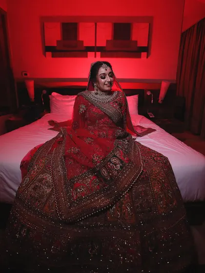 A full-length portrait of the bride seated on a bed, her magnificent lehenga spread out around her. The red ambient lighting creates a rich and intimate atmosphere.