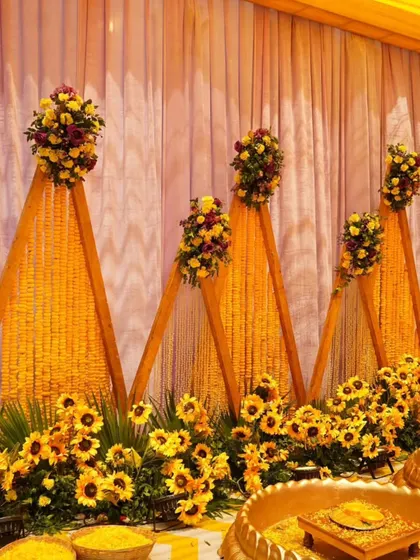 A unique Haldi backdrop design with triangular panels made of marigold strings. This setup includes a traditional urli and sunflower accents, perfect for an outdoor daytime event.