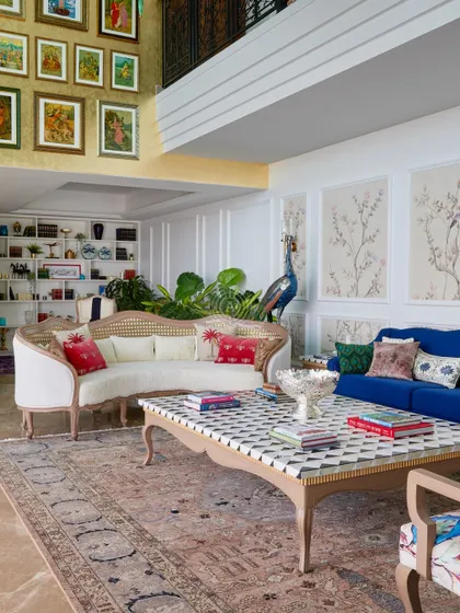 This living room, part of the 'Palace in the Sky' project, is designed for hosting. It features a double-height ceiling, a grand gallery wall, and a mix of custom furniture, including a classic cane sofa and a bold blue couch.