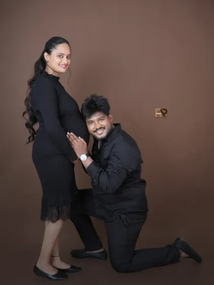 A playful and happy moment. The dad-to-be kneels to listen to the baby bump, and his joyful expression says it all. The coordinated black outfits create a sleek, modern look.